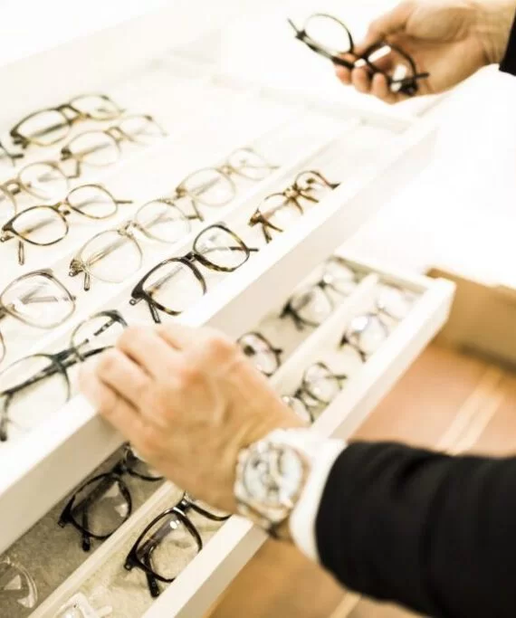Brillenregal bei Sattler Optik mit vielen Brillen, zwei Hände im Bild, die eine Brille herausnehmen